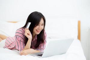 bela jovem asiática mentindo cartão de crédito de usuários com laptop, garota de conteúdo compras on-line e pagamento com notebook na cama no quarto, conceito de estilo de vida. foto