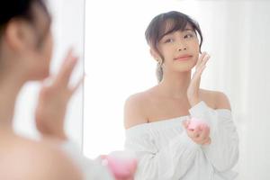 belo retrato jovem mulher asiática aplicando creme hidratante ou loção cosmética de cuidados com a pele no espelho do rosto, menina com tratamento facial, conceito de saúde e bem-estar. foto