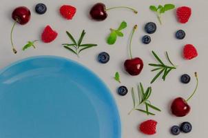 cerejas frescas, mirtilos, framboesas, hortelã e folha de alecrim na vista superior com placa azul e fundo de cor cinza pastel para o conceito de comida saudável. foto