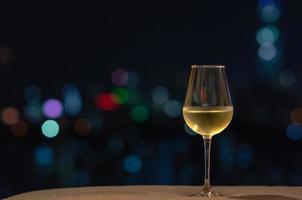 um copo com vinho branco coloca no sofá com luz colorida de bokeh da cidade. foto