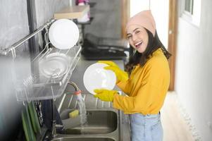 jovem mulher feliz usando luvas amarelas lavando pratos na cozinha em casa foto