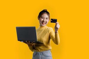 retrato de uma linda mulher asiática em uma camisa amarela, segurando um cartão de crédito e laptop. conceito de retrato usado para propaganda e sinalização, isolado sobre o fundo em branco, copie o espaço. foto