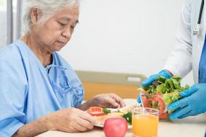 paciente idosa asiática ou idosa tomando café da manhã e comida saudável vegetal com esperança e feliz enquanto está sentado e com fome na cama no hospital. foto