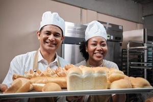 retrato de chefs profissionais de uniforme branco olhando para a câmera com um sorriso alegre e orgulhoso com bandeja de pão na cozinha. um amigo e parceiro de alimentos de panificação e ocupação diária de padaria fresca. foto