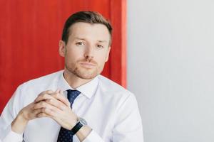 retrato de funcionário masculino bonito tem barba por fazer, mantém as mãos juntas, olha pensativamente para a câmera, contempla sobre o projeto de negócios, posa contra fundo vermelho e branco com espaço de cópia foto