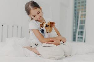 foto de menina bonita relaxada brinca com cachorro de pedigree, abraça o animal favorito, vestida de pijama casual, senta-se na cama, expressa amor ao animal de estimação, posa no quarto de luz. conceito de infância feliz