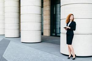 mulher bonita com cabelo encaracolado, pernas finas, vestindo fantasia preta e sapatos de salto alto, segurando o notebook nas mãos, organizando uma reunião com o parceiro de negócios em pé perto de um grande edifício de escritório foto