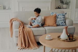 pequena adorável criança afro-americana sentada no sofá e brincando sozinha na sala de estar foto