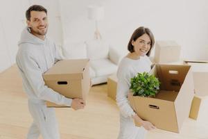 Proprietários positivos femininos e masculinos posam com pertences pessoais em caixas de papelão, mudam-se para o próprio apartamento, sorriem agradavelmente, posam na sala de estar branca. dia de mudança, planejamento, realocação foto