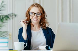 mulher encaracolada ruiva feliz trabalha como freelance, usa laptop, anota notas no bloco de notas, bebe café, usa óculos ópticos, está de bom humor, desenvolve empresa de negócios. conceito de trabalho foto