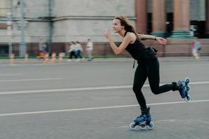 feriados e conceito de estilo de vida ativo. Slim saudável mulher europeia patins em alta velocidade photogaphed em movimento vestido com roupas pretas posa contra o fundo desfocado da rua na estrada. foto