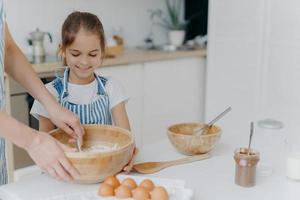 sorridente ajudante de menina segura tigela grande, olha como a mãe está misturando ovos com farinha, preparar bolo saboroso com chocolate, posar contra o interior aconchegante da cozinha em casa, preparar comida para toda a família foto