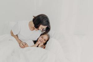 alegre mãe e filha se divertem juntas na cama, têm expressões felizes, desfrutam do conforto de roupas de cama macias, têm olhares felizes, passam a manhã no quarto. conceito de família, despertar e roupa de cama foto