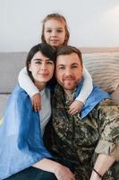 bandeira ucraniana está nos ombros. soldado de uniforme está em casa com sua esposa e filha foto