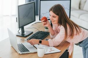 de pé com a maçã na mão e olhando para o laptop. corretora da bolsa feminina em roupas casuais está trabalhando no escritório pelo pc foto