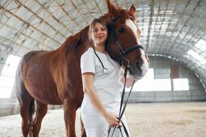 sorrindo e posando para a câmera. de pé juntos. médica de jaleco branco está com cavalo em um estábulo foto