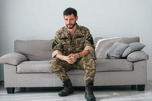 soldado de uniforme está dentro de casa na sala doméstica. sentado no sofá foto