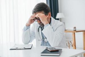 homem cansado, tem dor de cabeça. trabalhador médico profissional de jaleco branco está no escritório foto