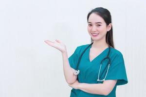 médica profissional asiática de uniforme verde fica e sorri enquanto aponta para o fundo branco. foto