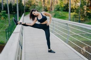 determinada esportista estica as pernas na ponte, usa roupas ativas, demonstra boa flexibilidade, posa ao ar livre contra o fundo da floresta. corredor de mulher se prepara para correr, tem treino de verão foto