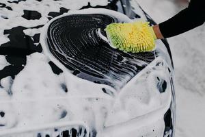 mão do homem com carro de limpeza de esfregão com xampu automático. espuma de sabão no capô preto. conceito de veículo. automóvel moderno coberto por espuma foto