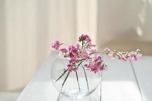 delicadas flores cor de rosa em vaso de vidro redondo. estética da primavera. foco seletivo. decoração de casa de estilo minimalista. mesa de madeira branca foto