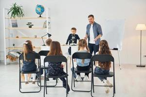 olhando para o menino com microscópio que está em cima da mesa. grupo de alunos de crianças em sala de aula na escola com professor foto