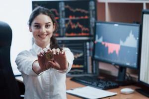 humor alegre. linda mulher segurando bitcoin nas mãos enquanto está sentado no escritório moderno com muitos monitores com gráficos foto