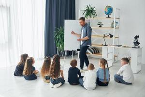 de pé e explicando a lição usando o quadro branco. grupo de alunos de crianças em sala de aula na escola com professor foto