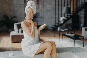 jovem relaxada linda jovem com maquiagem tem a pele limpa perfeita, aplica creme hidratante na loção para o corpo, posa envolta em toalha na sala de estar, parece feliz de lado. conceito de bem-estar foto