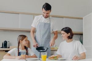 ocupado pai carinhoso prepara delicioso café da manhã para esposa e filha, usa avental, coloca ovos fritos no prato. alegre mãe e filho conversam enquanto se sentam à mesa da cozinha, fazem a refeição foto