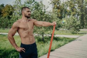 homem atlético musculoso tem treino com poses de banda de resistência elástica com corpo musculoso sem camisa, fica ao ar livre, concentrado na distância. desportista atleta tem treino no parque. estilo de vida saudável foto