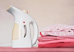 vaporizador de vestuário elétrico portátil e pilha de roupas infantis dobradas na tábua de engomar. limpeza e agregado familiar, conceito de tarefas. foto