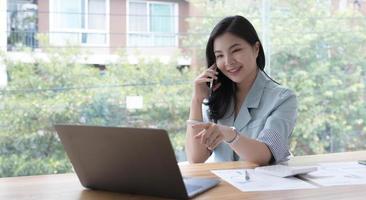 atraente jovem empresária asiática sorridente relaxando no escritório, trabalhando no computador portátil, falando no celular foto