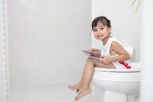 criança sentada no vaso sanitário segurando o tablet. conceito de smartphone viciado em criança foto