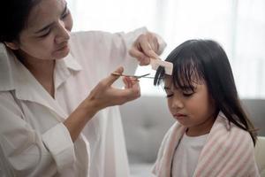 mãe asiática cortando cabelo para a filha na sala de estar em casa enquanto fica em casa segura do coronavírus covid-19 durante o bloqueio. conceito de auto-quarentena e distanciamento social.