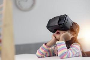 criança com fone de ouvido de realidade virtual sentado atrás da mesa dentro de casa em casa foto