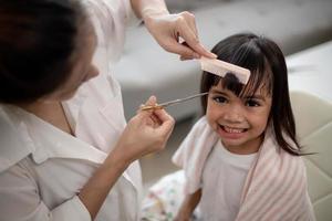 mãe asiática cortando cabelo para a filha na sala de estar em casa enquanto fica em casa segura do coronavírus covid-19 durante o bloqueio. conceito de auto-quarentena e distanciamento social. foto