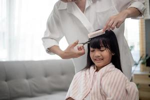 mãe asiática cortando cabelo para a filha na sala de estar em casa enquanto fica em casa segura do coronavírus covid-19 durante o bloqueio. conceito de auto-quarentena e distanciamento social. foto