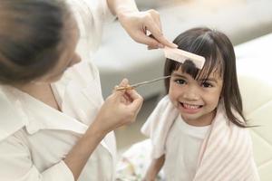 mãe asiática cortando cabelo para a filha na sala de estar em casa enquanto fica em casa segura do coronavírus covid-19 durante o bloqueio. conceito de auto-quarentena e distanciamento social. foto