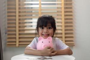 menina asiática economizando dinheiro em um cofrinho, aprendendo sobre economia, criança economizando dinheiro para educação futura. conceito de dinheiro, finanças, seguro e pessoas foto