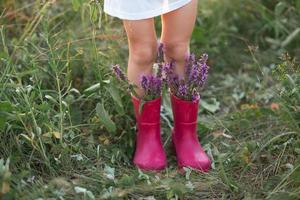 botas de borracha vermelha nos pés de uma garota com um buquê de flores silvestres. flores em uma bota, horário de verão. verão, liberdade, natureza, campo, grama verde no campo foto