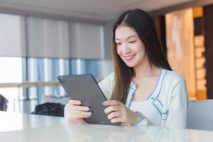 mulher profissional asiática jovem adulta bonita com cabelos compridos na camisa branca detém o tablet dela sorrindo alegremente enquanto ela se senta na cadeira no escritório do local de trabalho com edifício envidraçado como pano de fundo. foto