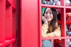 mulher bonita asiática de cabelos compridos vestindo um manto amarelo e sorriso feliz em pé na cabine telefônica vermelha no tema de celebrar o natal e feliz ano novo foto