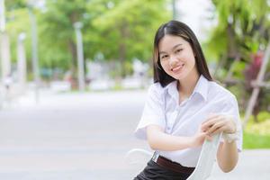 retrato de estudante tailandês adulto em uniforme de estudante universitário. menina bonita asiática sentado sorrindo alegremente na universidade ao ar livre com um fundo de árvores de jardim ao ar livre. foto