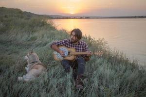 folk ou country guitarrista na floresta com cachorro, homem na floresta, fogueira e fundo por do sol foto