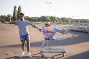 casal jovem feliz andando de carrinho no estacionamento vazio do shopping, amigo hipster se divertindo durante as compras, casal apaixonado andando no carrinho de compras foto