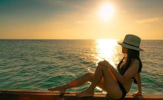 sexy e relaxa a mulher usa biquíni preto com chapéu de palha sente-se na viga de madeira perto da praia de areia ao pôr do sol. garota aproveite as férias na praia do paraíso tropical nas férias de verão. viagens de férias. Ritmo de verão. foto