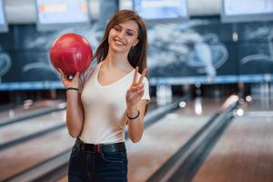 jovem alegre em roupas casuais, segurando uma bola de boliche vermelha no clube foto