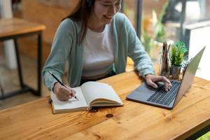 jovem estudante asiática usa fones de ouvido escrevendo no notebook para estudar o idioma online assistir e ouvir o palestrante, webinar via videochamada e-learning em casa foto